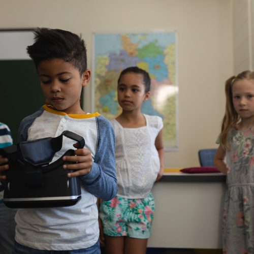 Front view of schoolboy holding and looking at virtual reality headset with his classmates who look him behind  in classroom of elementary school