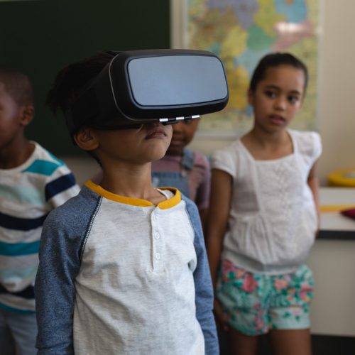 Front view of schoolboy using virtual reality headset whit his classmates behind him in classroom of elementary school