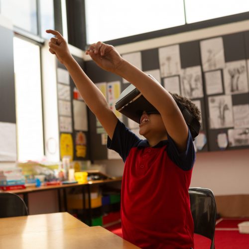 Side view of cute mixed-race schoolboy using virtual reality headset with hands in the air at desk in a classroom at elementary school